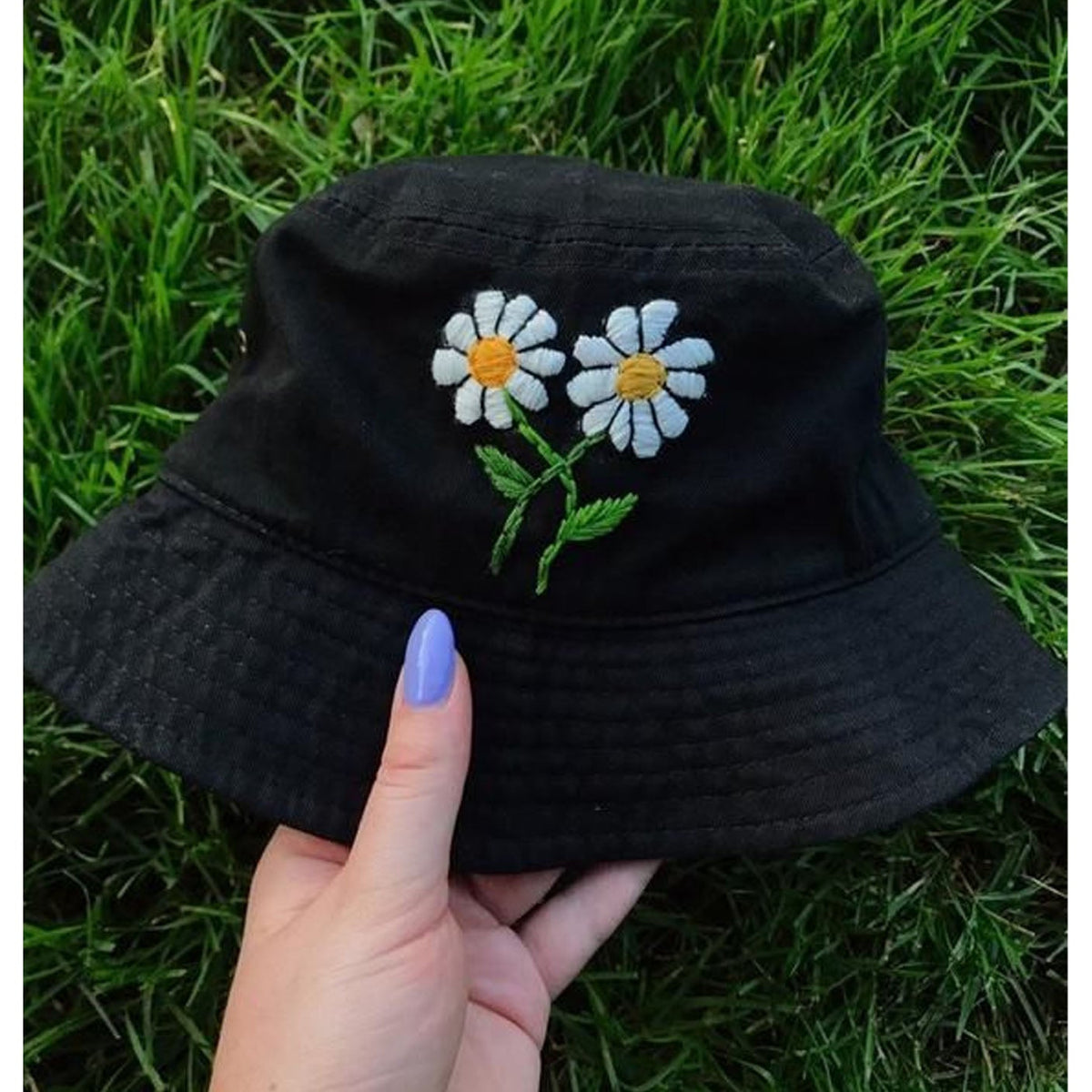 A black bucket hat with embroidered white daisies featuring yellow centers and green stems, held by a hand with purple nail polish against a background of green grass.