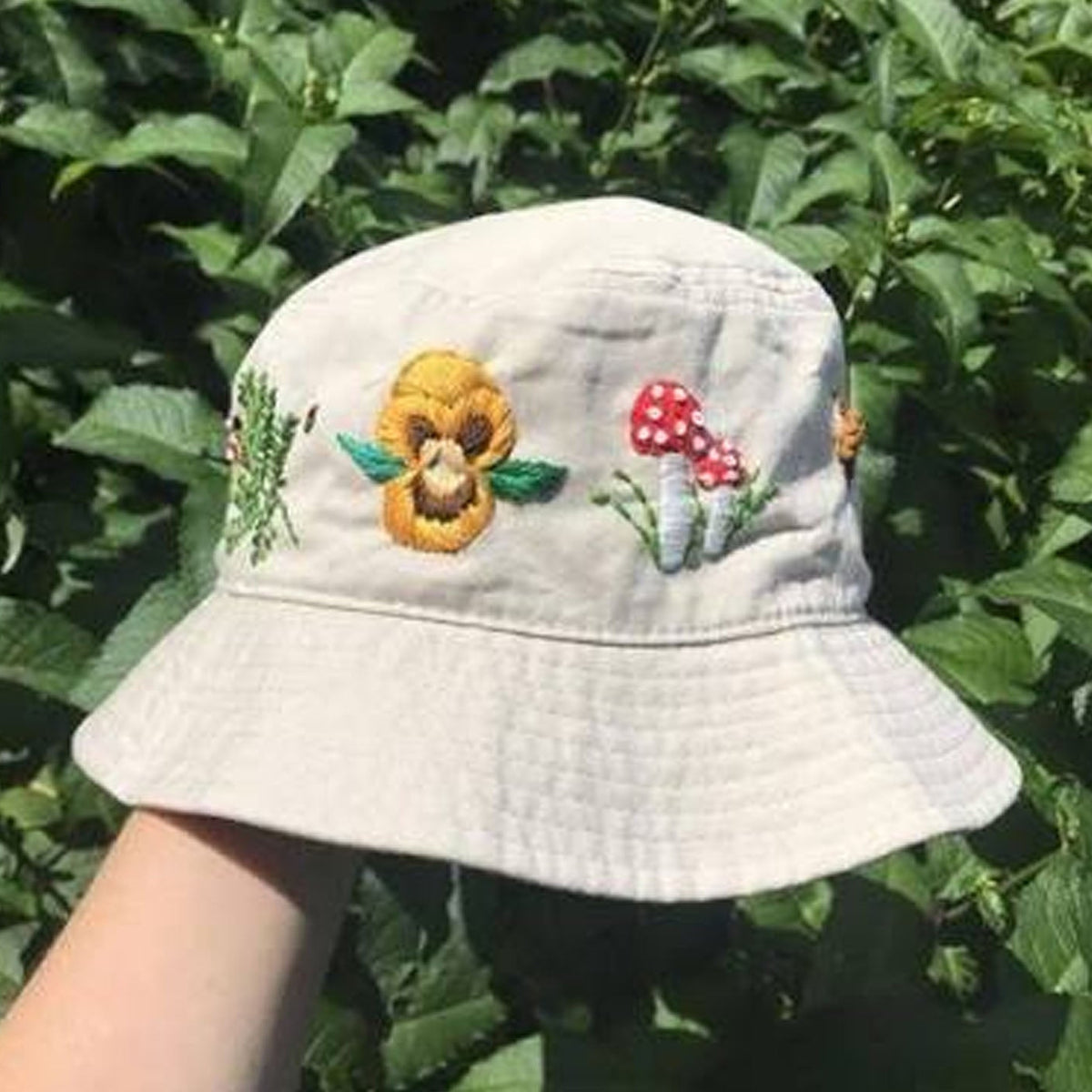 A light beige bucket hat with embroidered wildflowers and mushrooms, held by a hand against a background of green leaves.