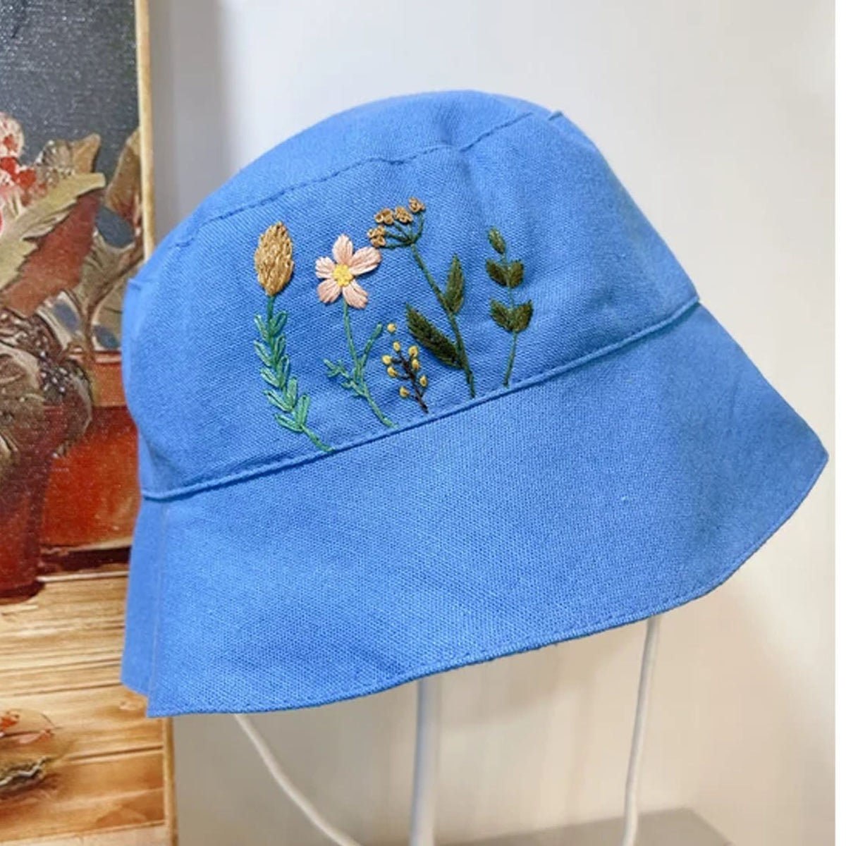 A blue bucket hat with colorful floral embroidery on the front, displayed on a white stand against a neutral indoor background.