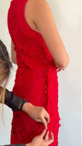 A woman wearing a bright red sleeveless dress is shown from the side, with another person adjusting the back of the dress against a plain light background.
