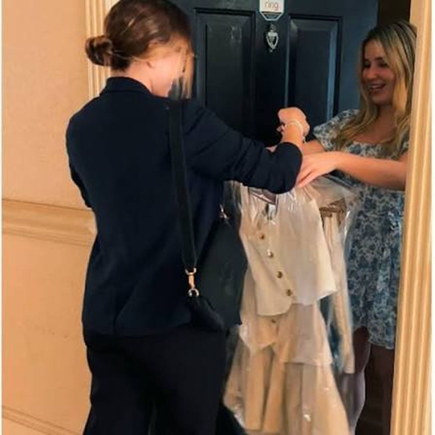 A woman in a navy blazer and black pants holds up a white and beige tiered dress in a clear plastic garment bag, showing it to another woman standing in a doorway.