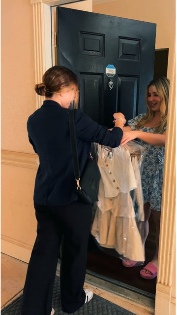A woman in a navy blazer and black pants receives a cream-colored dress from another woman standing in a doorway.