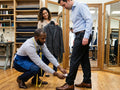 A man in a light blue dress shirt and dark trousers stands while a tailor crouches to measure his leg with a yellow tape measure. A woman holds up a dark gray suit jacket in a well-lit tailoring shop with wood floors and mirrors.