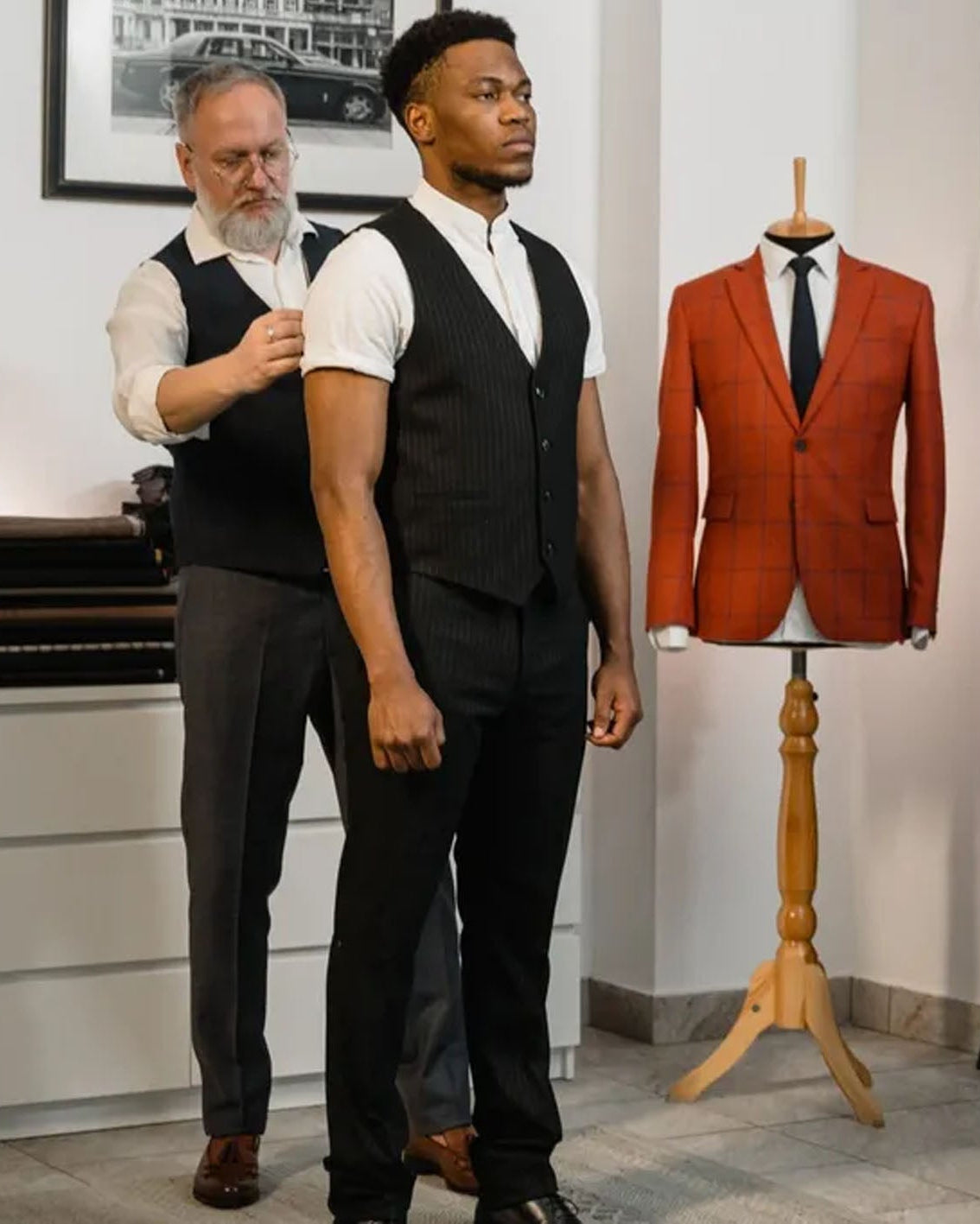 A man in a black pinstripe vest, white shirt, and black trousers stands indoors while another man adjusts his vest. A mannequin wearing a rust-orange pinstripe suit jacket is displayed on a wooden stand to the right.
