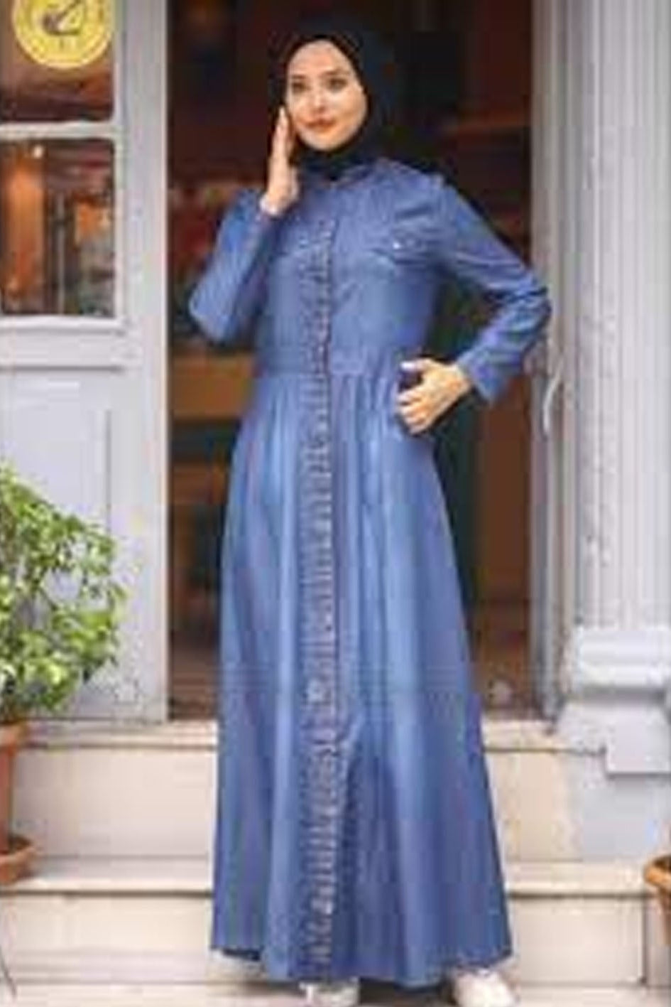 Woman wearing a long blue dress with buttons, standing in front of a door.