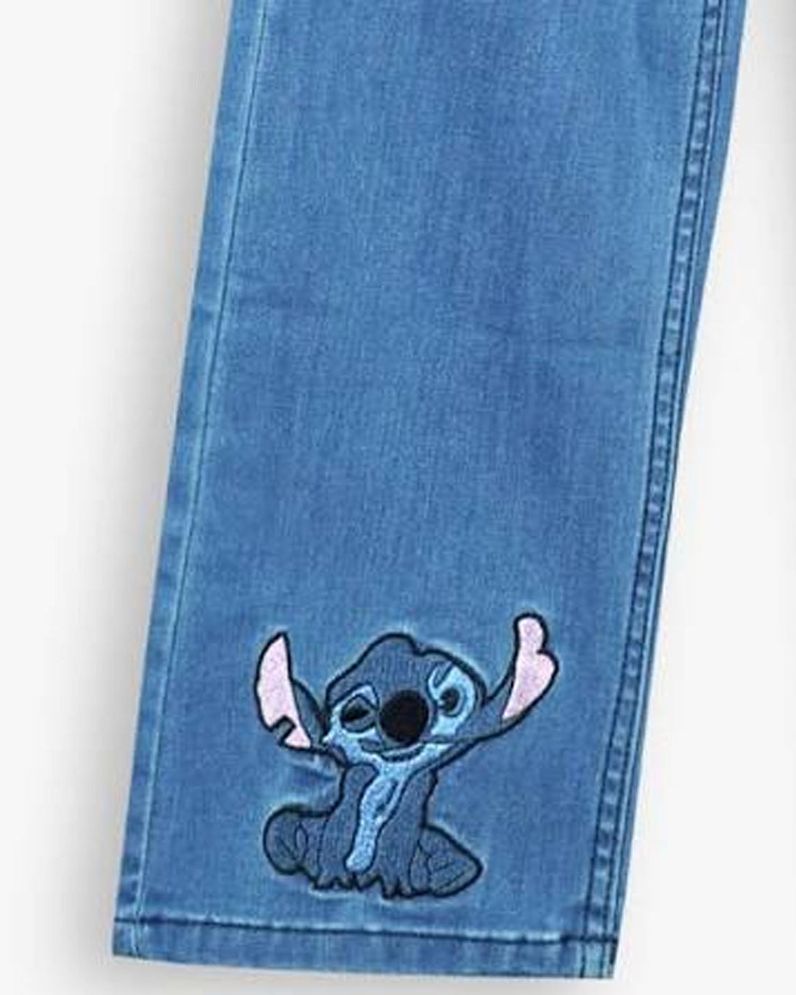 A close-up of a blue denim jeans leg featuring an embroidered patch of the cartoon character Stitch on the lower front.