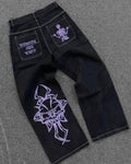 Black denim jeans with purple graphic prints, laid flat on a gray asphalt surface. The back pocket features a purple skeleton graphic and white text, while the front leg has a large purple gothic-style graphic.