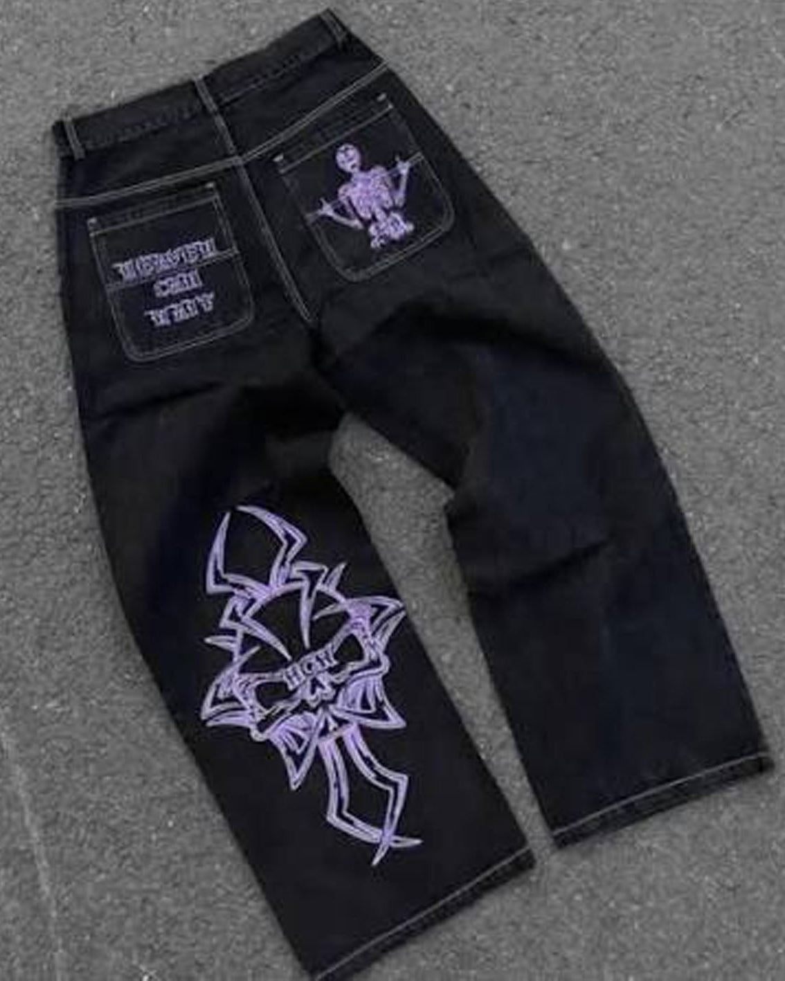 Black denim jeans with purple graphic prints, laid flat on a gray asphalt surface. The back pocket features a purple skeleton graphic and white text, while the front leg has a large purple gothic-style graphic.