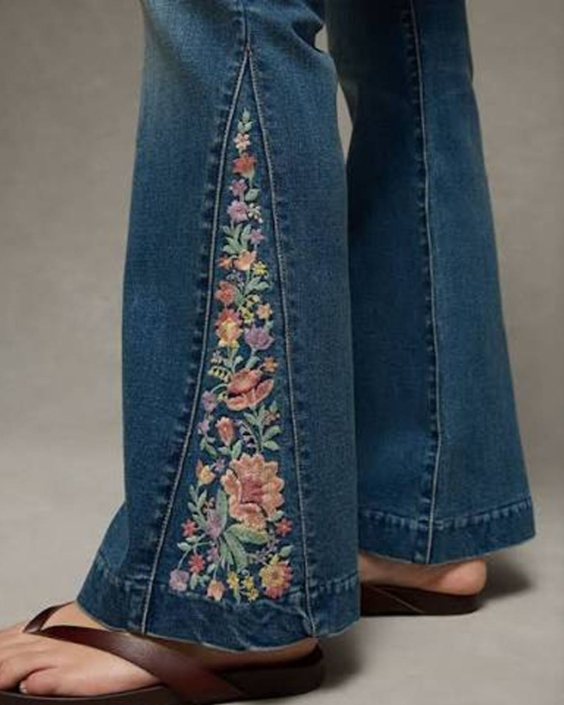 Close-up of a person's legs wearing blue denim flare jeans with colorful floral embroidery down the front of each leg, paired with brown sandals against a neutral gray background.