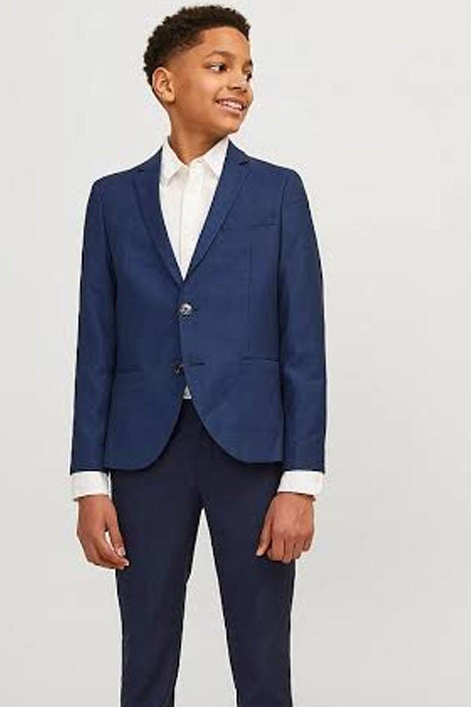 A young boy modeling a navy blue two-piece suit with a white collared shirt, standing against a light gray studio background.