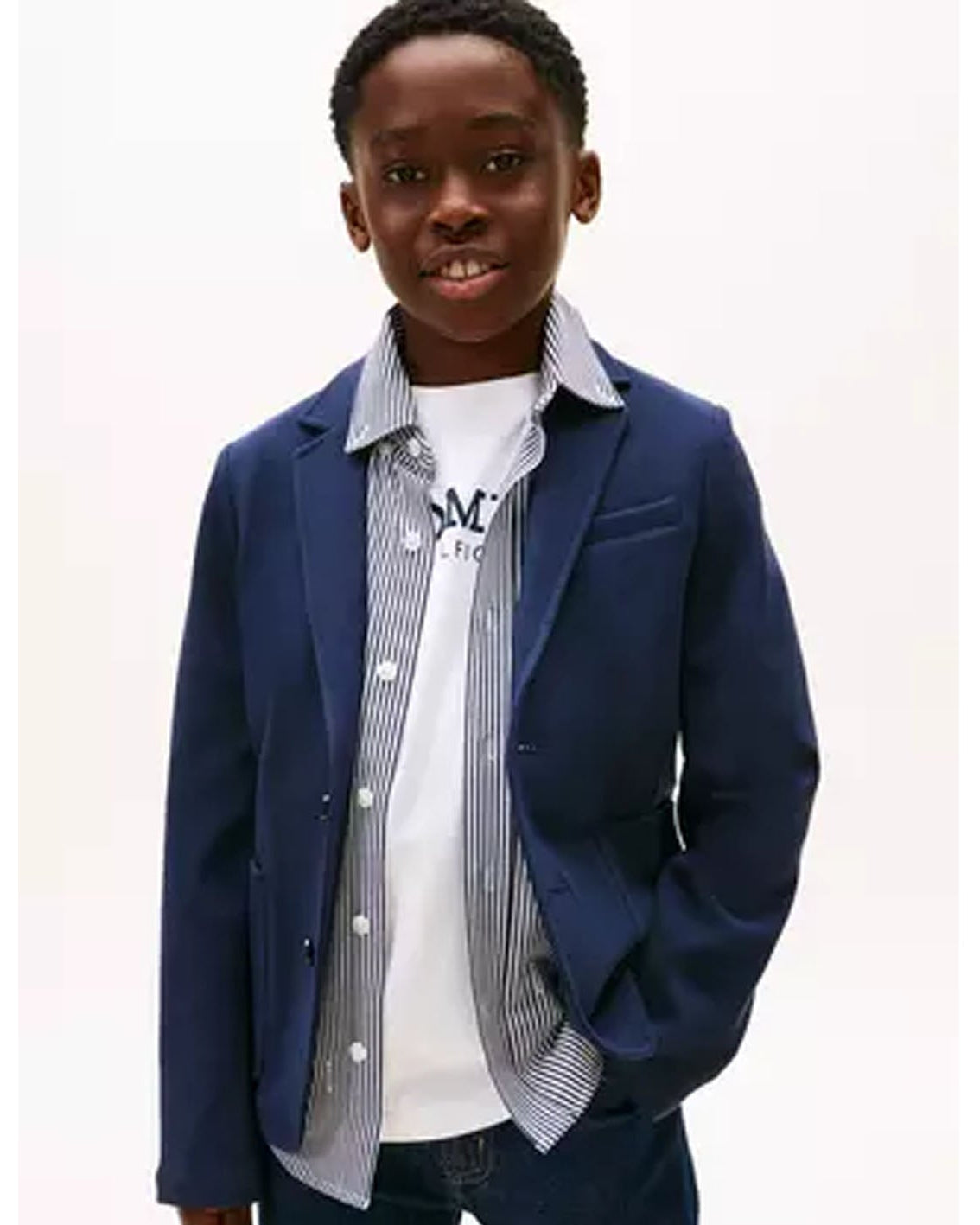 A young boy modeling a navy blue blazer over a white t-shirt and a blue-and-white striped button-up shirt, set against a plain white background.