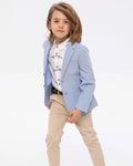 A young boy modeling a light blue blazer over a white patterned shirt and beige trousers, standing against a plain white studio background.