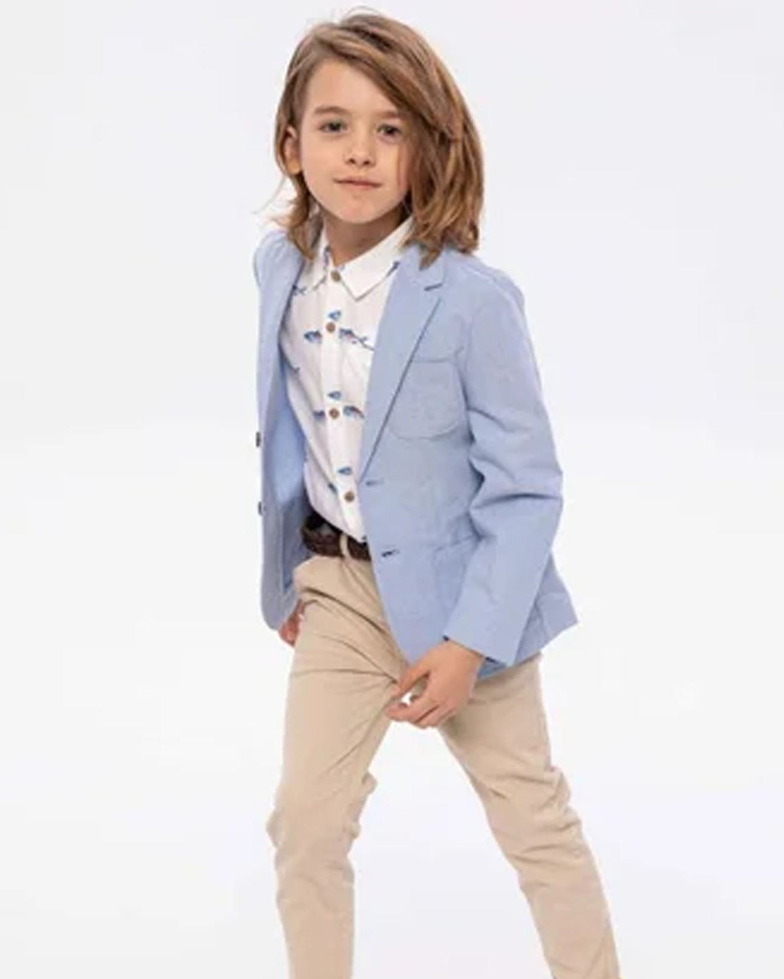 A young boy modeling a light blue blazer over a white patterned shirt and beige trousers, standing against a plain white studio background.