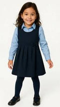 Young girl wearing a navy pinafore dress over a light blue shirt on a white background