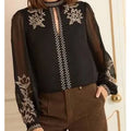 Person wearing a black blouse with floral embroidery and brown pants indoors.