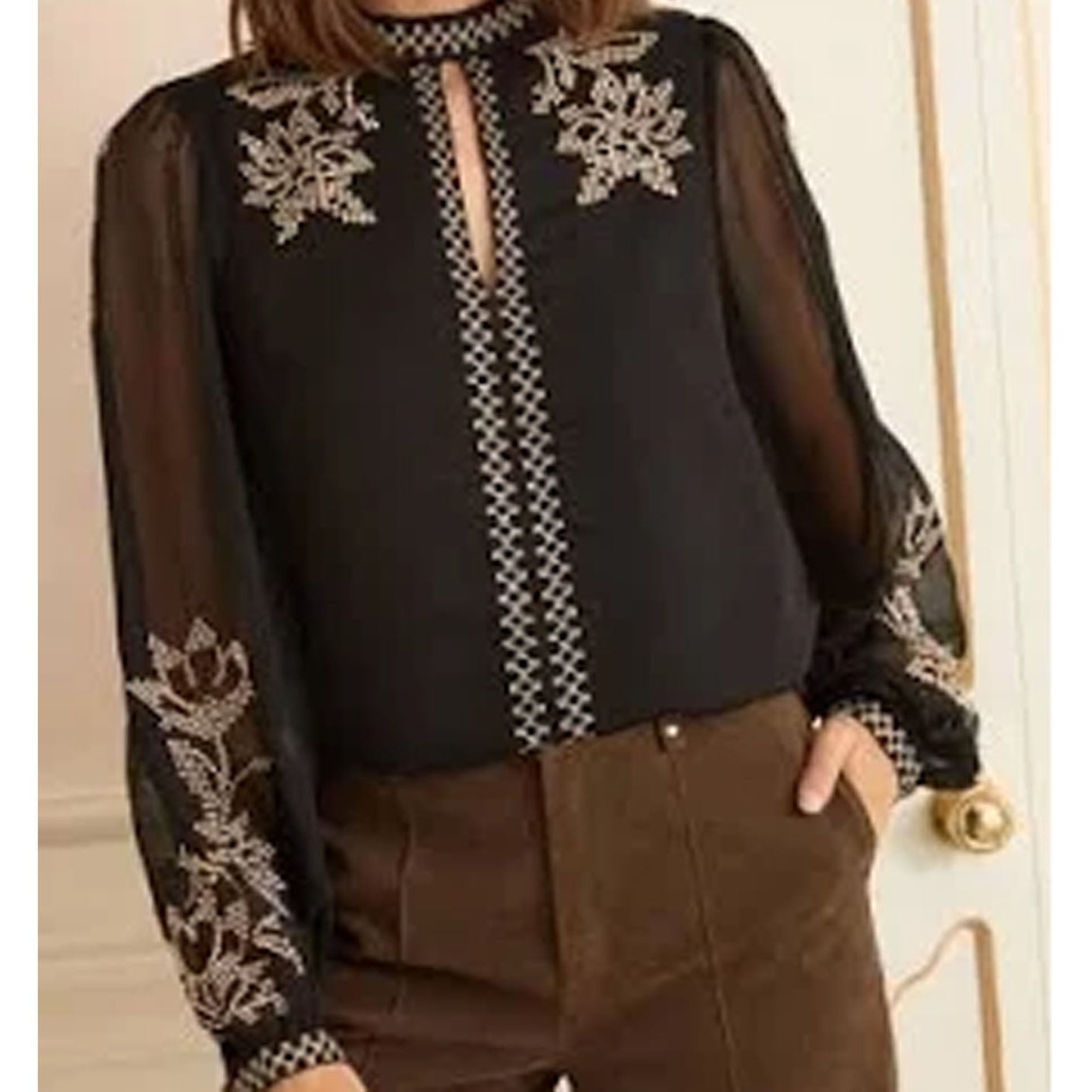 Person wearing a black blouse with floral embroidery and brown pants indoors.