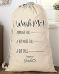 A beige fabric laundry bag with black printed text reading 'Wash Me! ALMOST FULL A BIT MORE FULL A BIT FULL Property of: Charlotte' in a home setting.