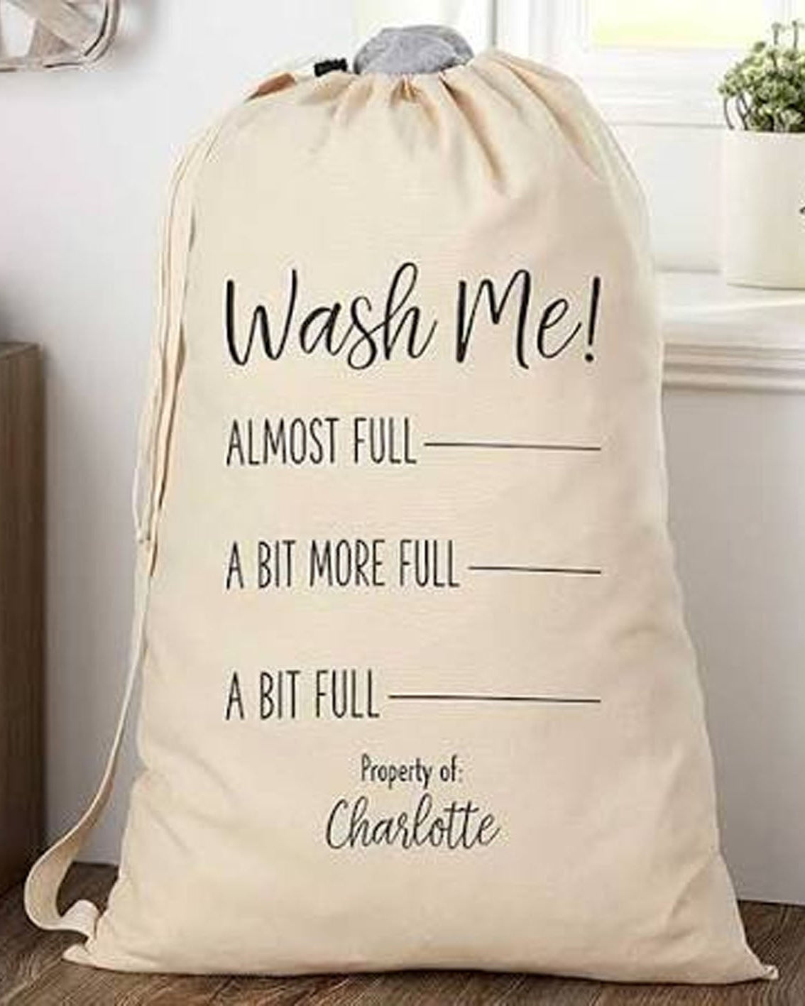 A beige fabric laundry bag with black printed text reading 'Wash Me! ALMOST FULL A BIT MORE FULL A BIT FULL Property of: Charlotte' in a home setting.