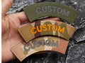 Three curved embroidered patches with the word 'CUSTOM' are held in a hand. The top patch is olive green with gray lettering, the middle patch has a tan and brown camouflage pattern with orange lettering, and the bottom patch is a tan and brown camouflage pattern with gray lettering.