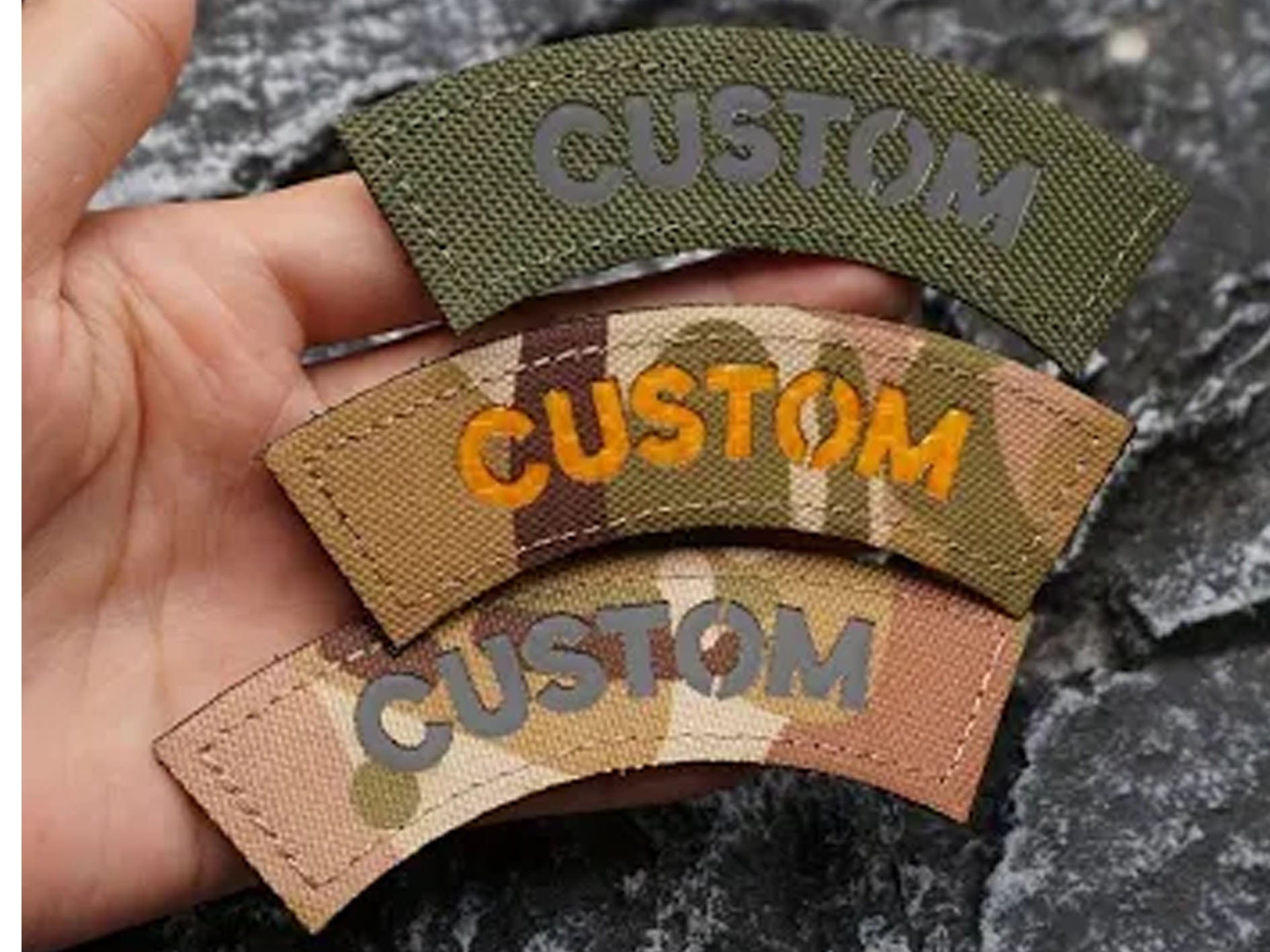 Three curved embroidered patches with the word 'CUSTOM' are held in a hand. The top patch is olive green with gray lettering, the middle patch has a tan and brown camouflage pattern with orange lettering, and the bottom patch is a tan and brown camouflage pattern with gray lettering.