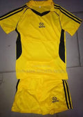 A yellow and black athletic soccer kit consisting of a short-sleeve jersey and matching shorts, laid flat on a gray tiled floor. The jersey features black shoulder stripes, black side panels, and a black V-neck collar, with a black 'sports' logo on the chest.