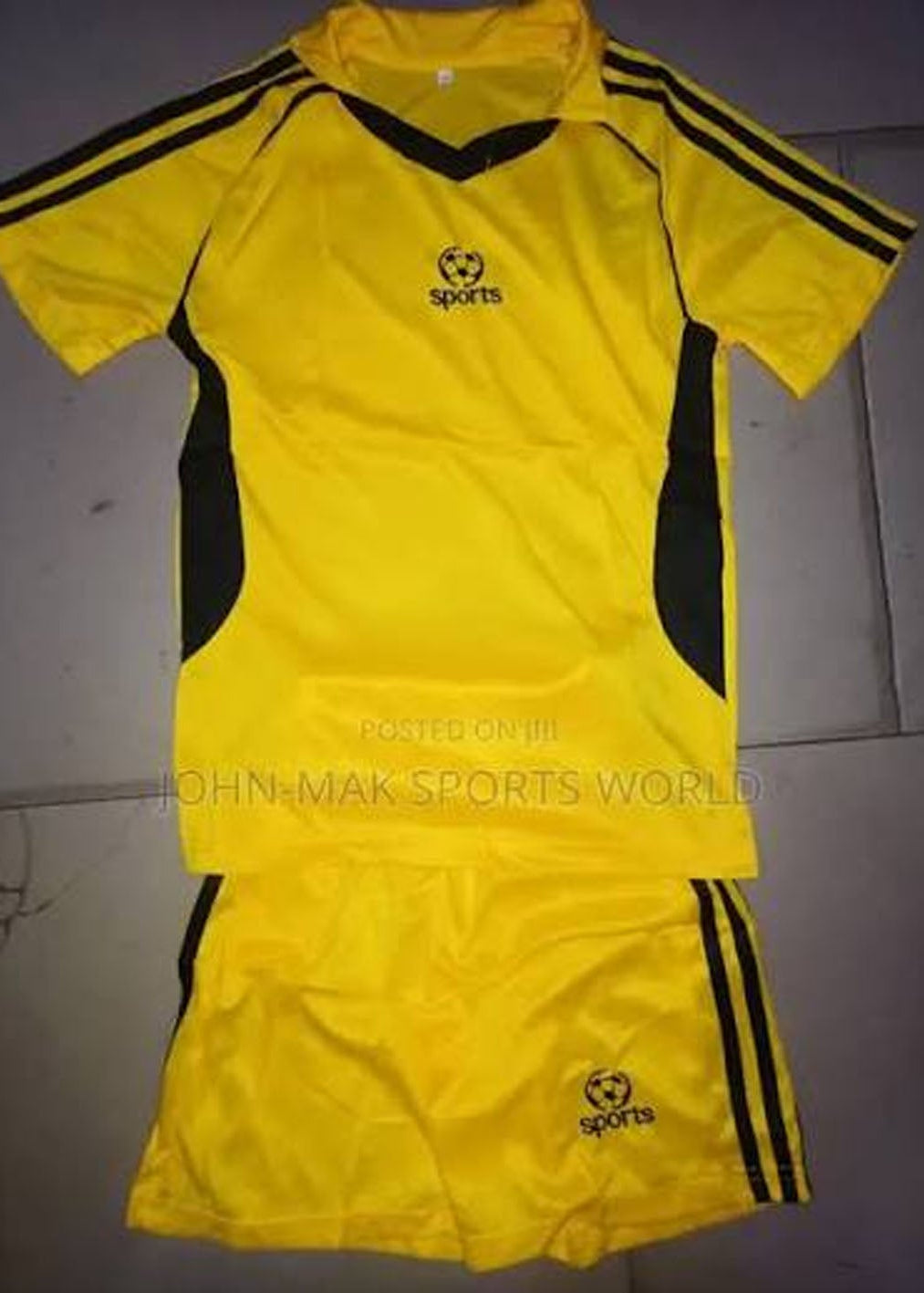 A yellow and black athletic soccer kit consisting of a short-sleeve jersey and matching shorts, laid flat on a gray tiled floor. The jersey features black shoulder stripes, black side panels, and a black V-neck collar, with a black 'sports' logo on the chest.