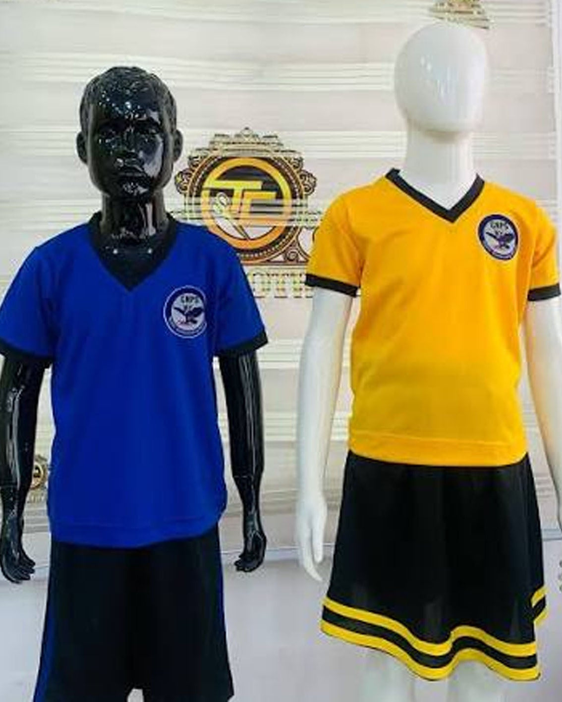 Two children's sports jerseys displayed on mannequins: a royal blue V-neck jersey with black shorts on a black mannequin, and a bright yellow V-neck jersey with black trim and a black skirt with yellow stripes on a white mannequin. A decorative wall plaque is behind them.