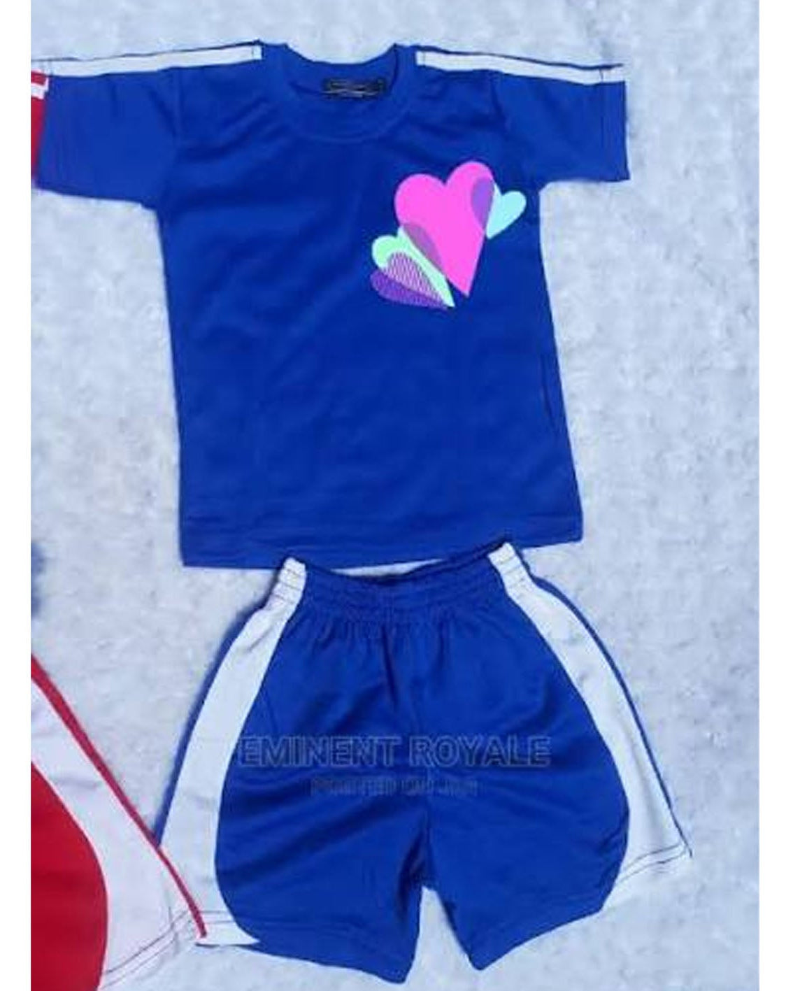 A kids' two-piece athletic set featuring a royal blue short-sleeve t-shirt with white shoulder stripes and a pink, purple, and green heart graphic on the chest, paired with matching blue shorts that have white side stripes.