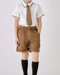 A child wearing a school uniform set consisting of a white short-sleeve collared shirt with brown trim, a brown tie, brown shorts, and black knee-high socks, photographed against a light gray studio background.