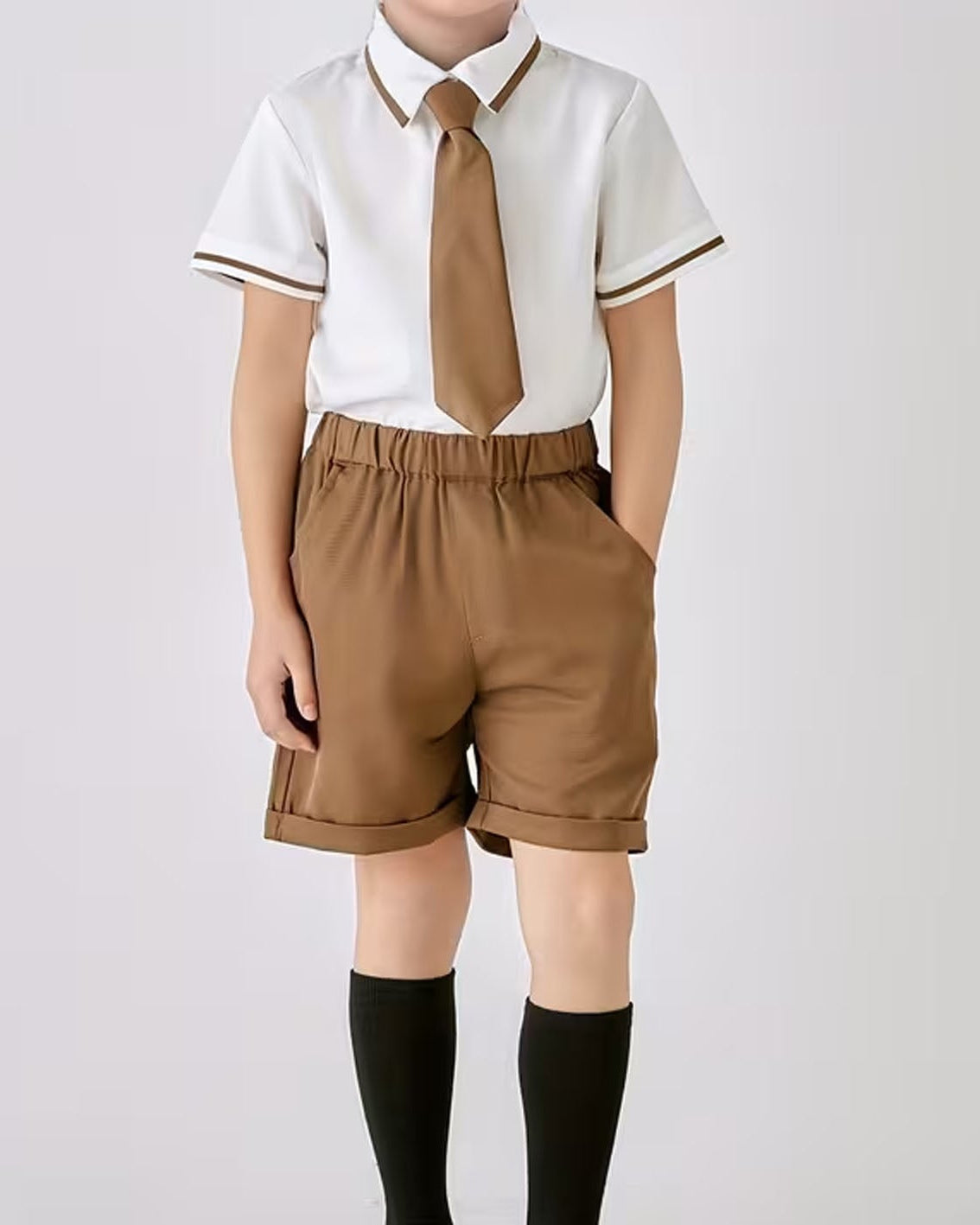 A child wearing a school uniform set consisting of a white short-sleeve collared shirt with brown trim, a brown tie, brown shorts, and black knee-high socks, photographed against a light gray studio background.