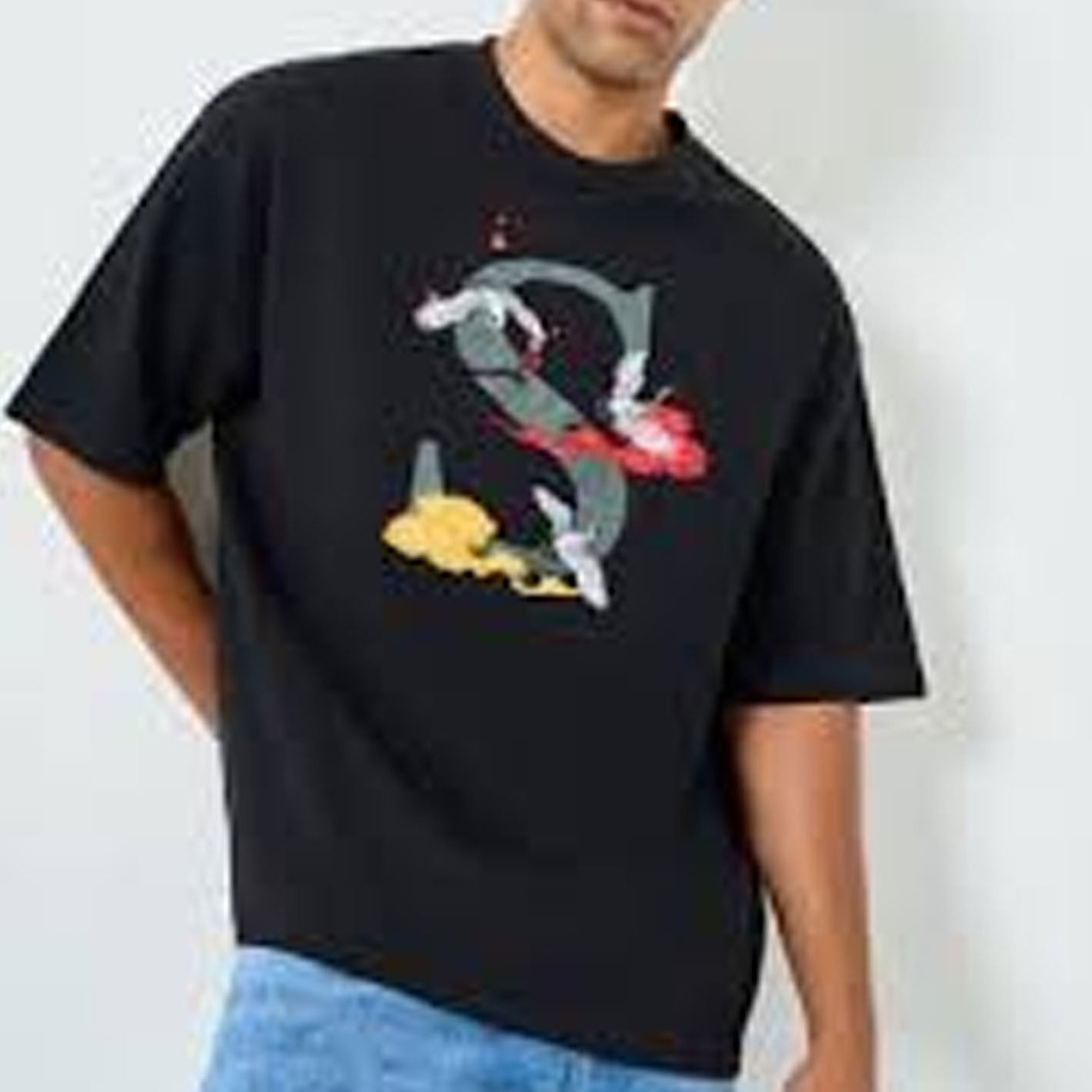 A man wearing a black short-sleeve t-shirt with a colorful graphic of a cartoon cat and mouse on the front, paired with blue jeans, against a plain light gray background.