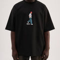 A person wearing an oversized black t-shirt with a central graphic of a walking figure in a red cap and blue pants, with the word 'now' printed below.