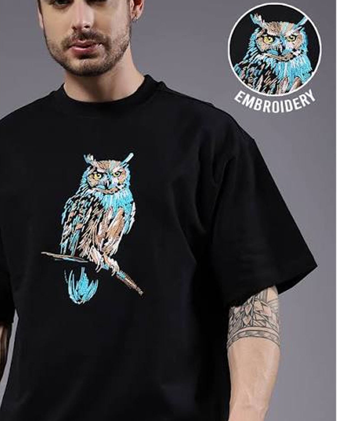 A man wearing a black crewneck t-shirt with a detailed graphic of an owl perched on a branch in blue, white, and brown tones. A circular badge in the top right corner highlights the embroidery detail of the graphic.