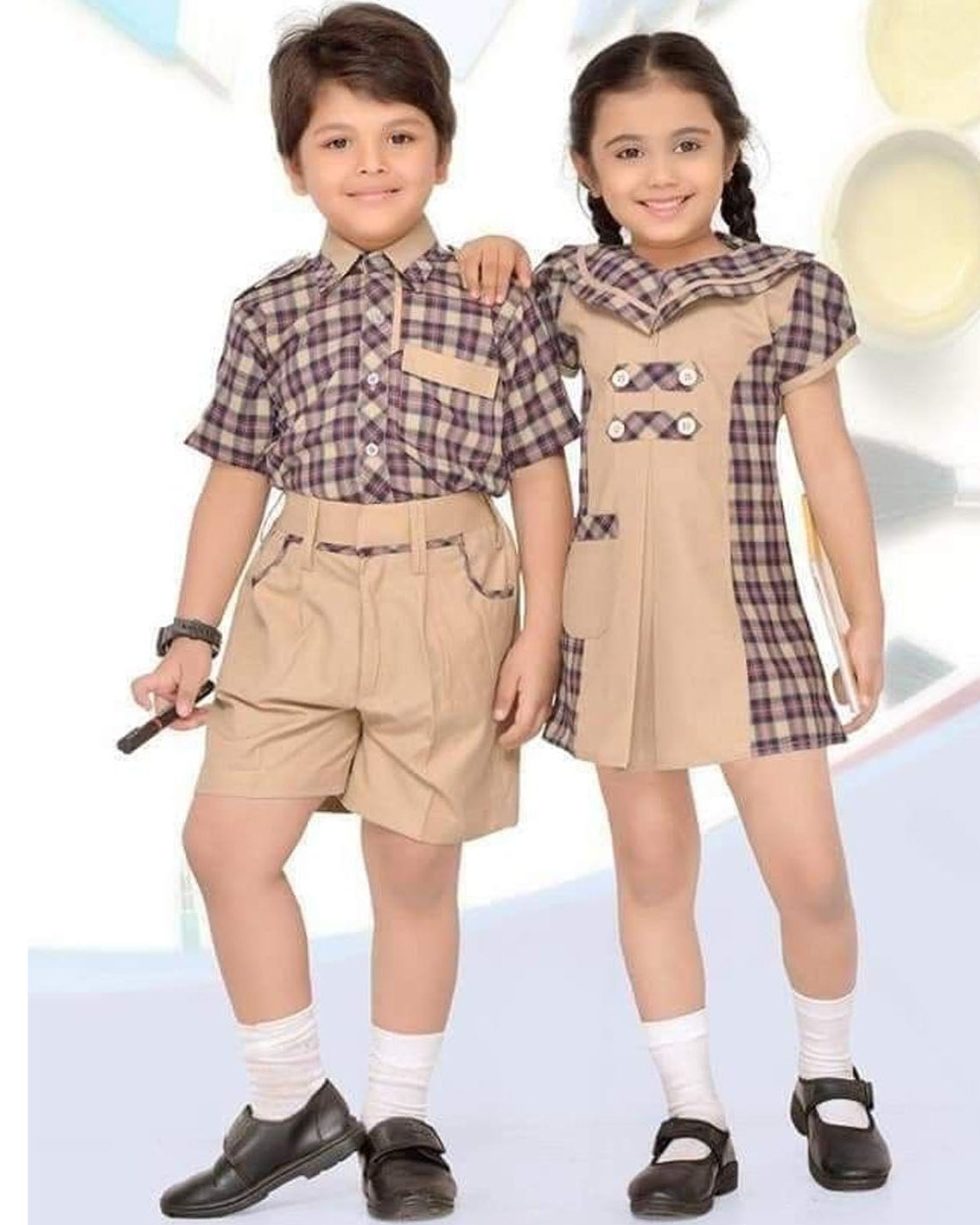 A boy and a girl modeling matching school uniforms. The boy wears a short-sleeve plaid button-up shirt in purple and beige with khaki shorts, white socks, and black shoes.