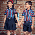 Two children wearing matching striped school uniforms holding hands in front of a brick wall. The girl wears a navy pleated skirt and the boy wears navy shorts, both in red, blue, and white striped shirts with navy blazers and belts.