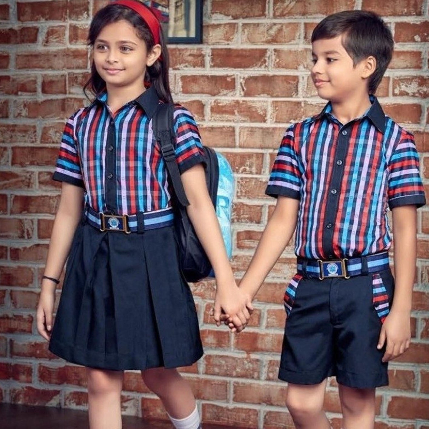 Two children wearing matching striped school uniforms holding hands in front of a brick wall. The girl wears a navy pleated skirt and the boy wears navy shorts, both in red, blue, and white striped shirts with navy blazers and belts.