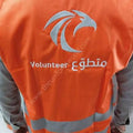 An orange high-visibility volunteer vest with reflective silver stripes and a silver eagle logo, worn over gray sleeves. The back of the vest features the text 'Volunteer' and Arabic script.