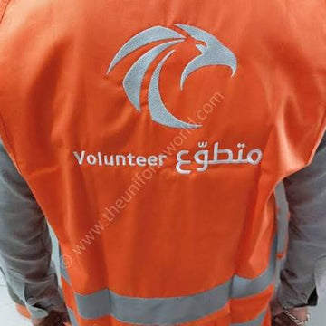An orange high-visibility volunteer vest with reflective silver stripes and a silver eagle logo, worn over gray sleeves. The back of the vest features the text 'Volunteer' and Arabic script.
