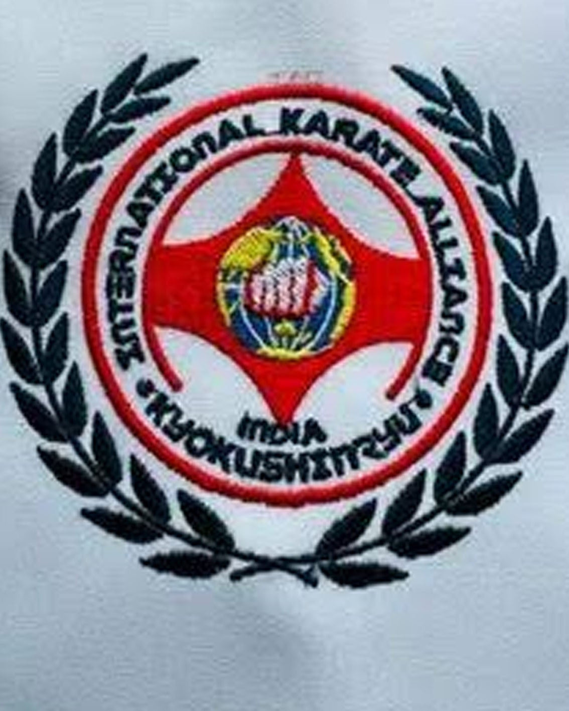 Close-up of an embroidered circular logo on a white fabric, featuring a red ring with black text reading 'International Karate Alliance India' around a central emblem with a fist and globe, surrounded by a black laurel wreath.