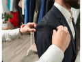 A man wearing a dark navy suit jacket and white dress shirt has another person adjusting the shoulder fit with a pin cushion and sewing pins visible in the background of a clothing store.