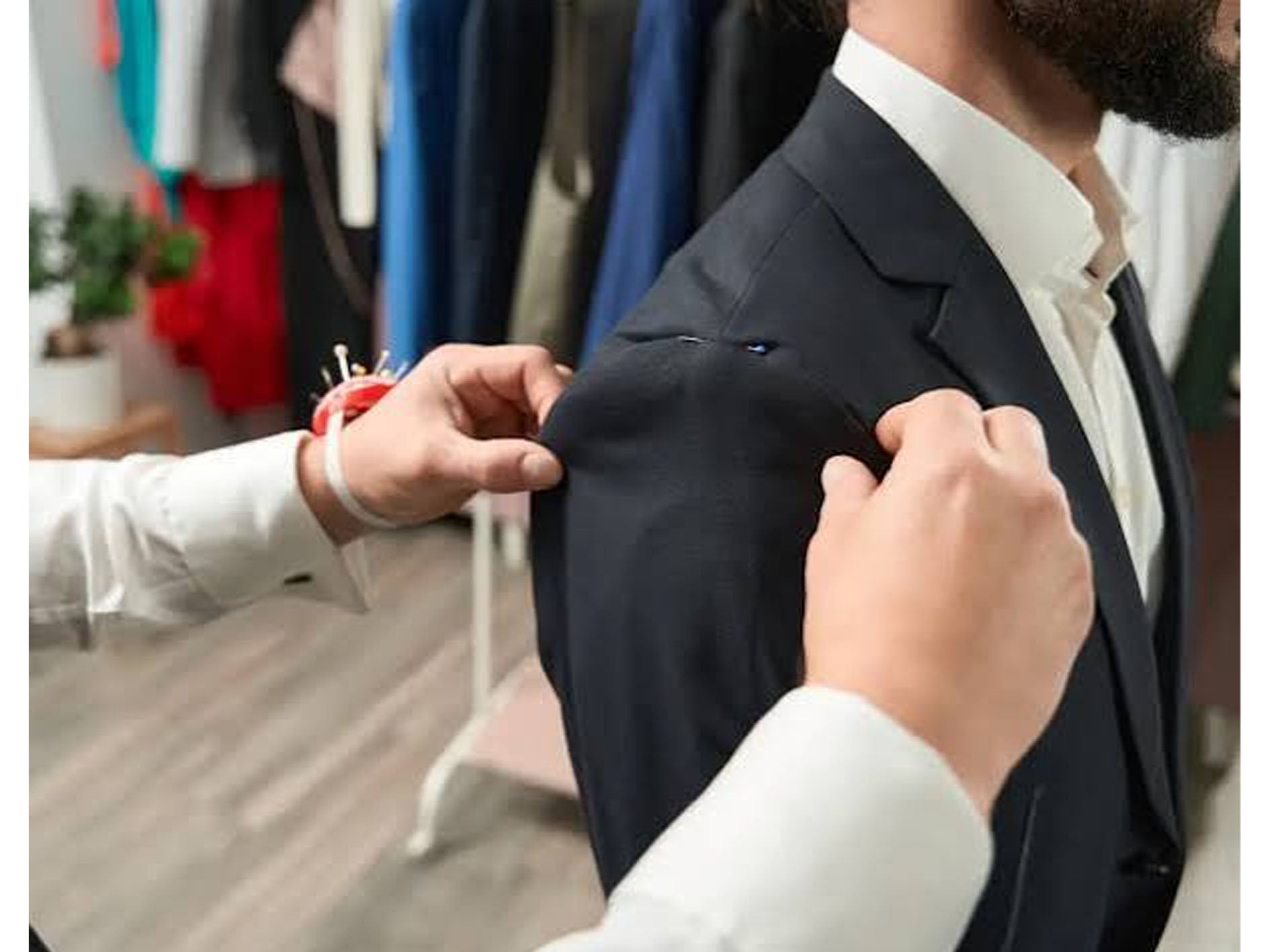 A man wearing a dark navy suit jacket and white dress shirt has another person adjusting the shoulder fit with a pin cushion and sewing pins visible in the background of a clothing store.