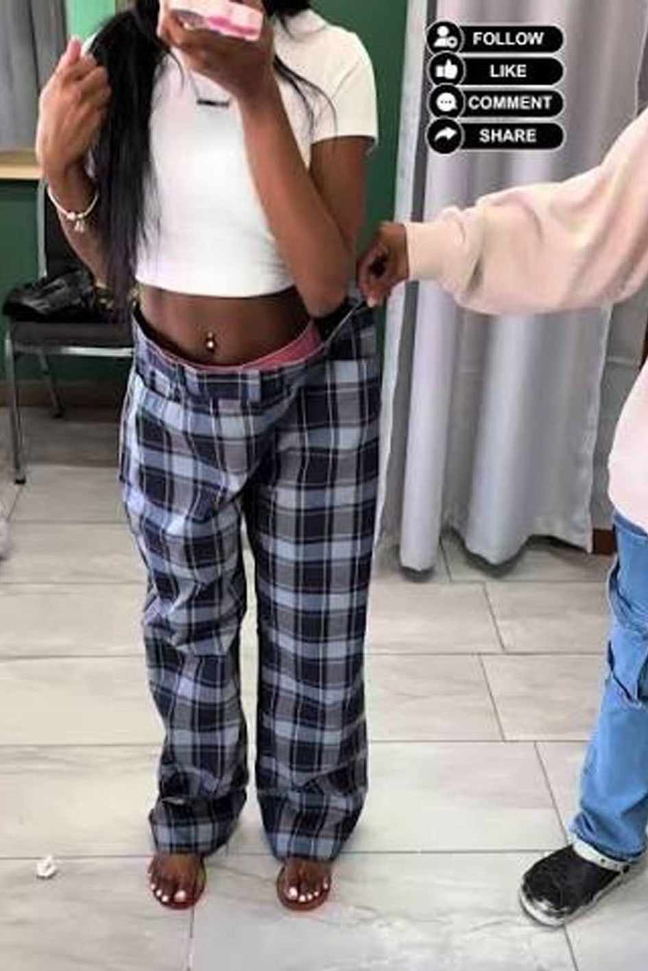 A person taking a mirror selfie indoors wearing white cropped shorts with a pink waistband and loose black-and-white plaid pants. Another person's arm is visible adjusting the shorts.