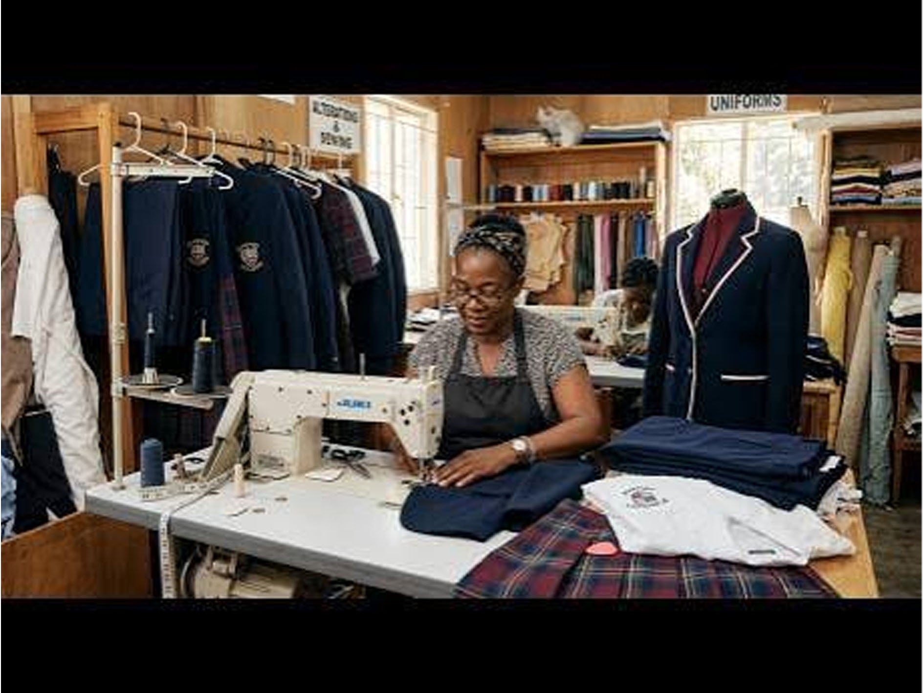 A woman sews navy blue uniforms at a white sewing machine inside a wooden tailoring workshop. Jackets and folded fabric are visible on racks and tables, with a white shirt laid out on the table in the foreground.