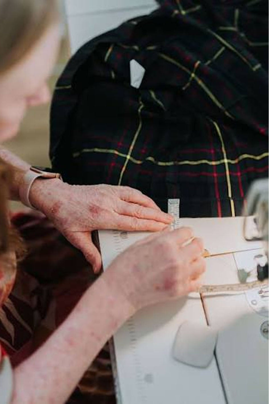 A close-up lifestyle shot of a person's hands measuring a black tartan plaid fabric with a ruler at a sewing machine.