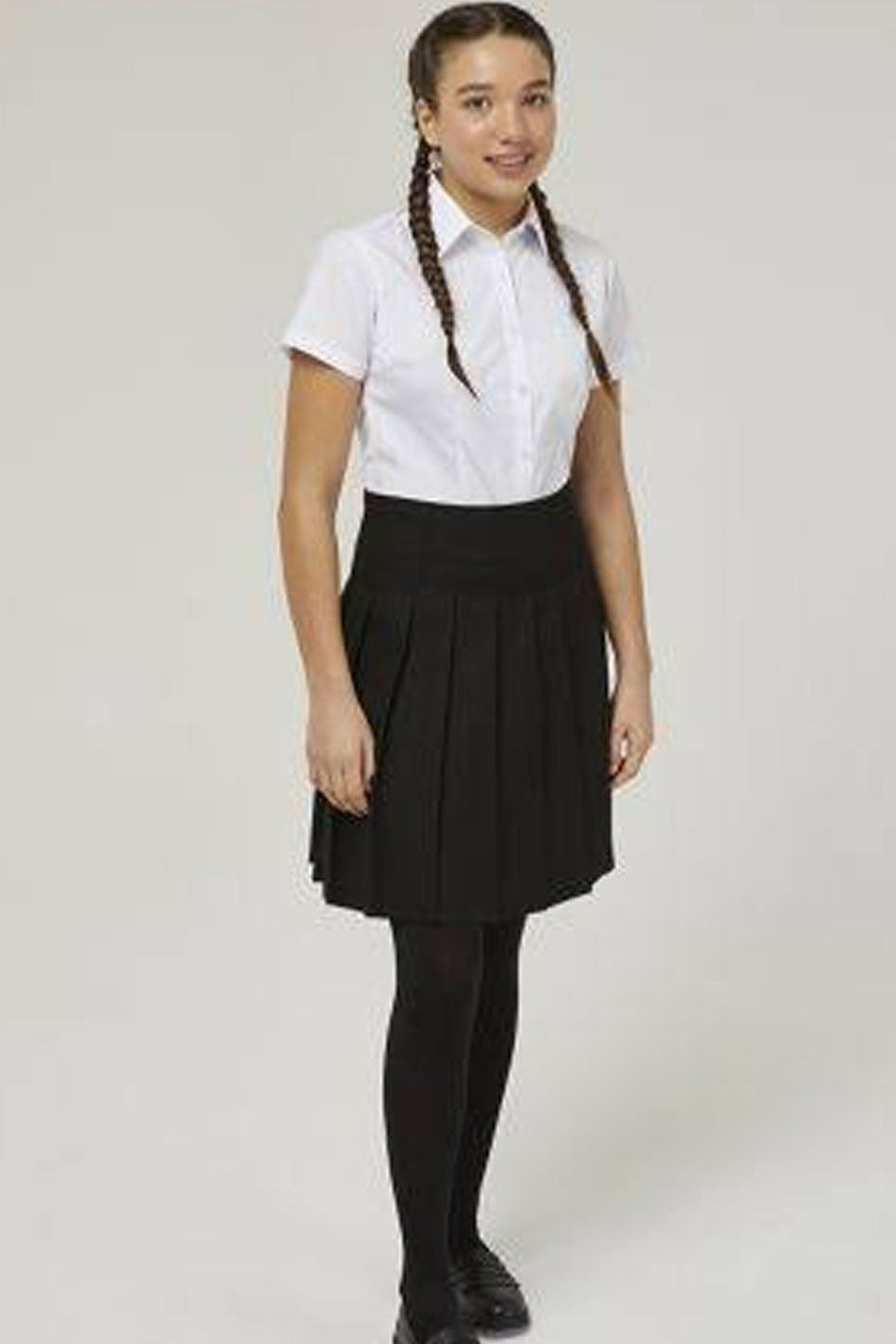 A girl modeling a school uniform consisting of a white short-sleeve button-up shirt, a black pleated skirt, black tights, and black shoes, standing against a light gray studio background.