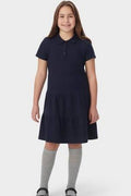 A young girl modeling a short-sleeve navy blue dress with a tiered skirt, paired with light gray knee-high socks and black shoes, against a light gray background.