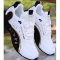 A pair of white low-top sneakers with black side panels, gold metallic accents, and white laces, shown against a blurred outdoor background.
