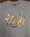 A white short-sleeve t-shirt featuring a metallic gold foil graphic that reads 'Bride' in cursive, decorated with small hearts and a ring motif, shown on a hanger.