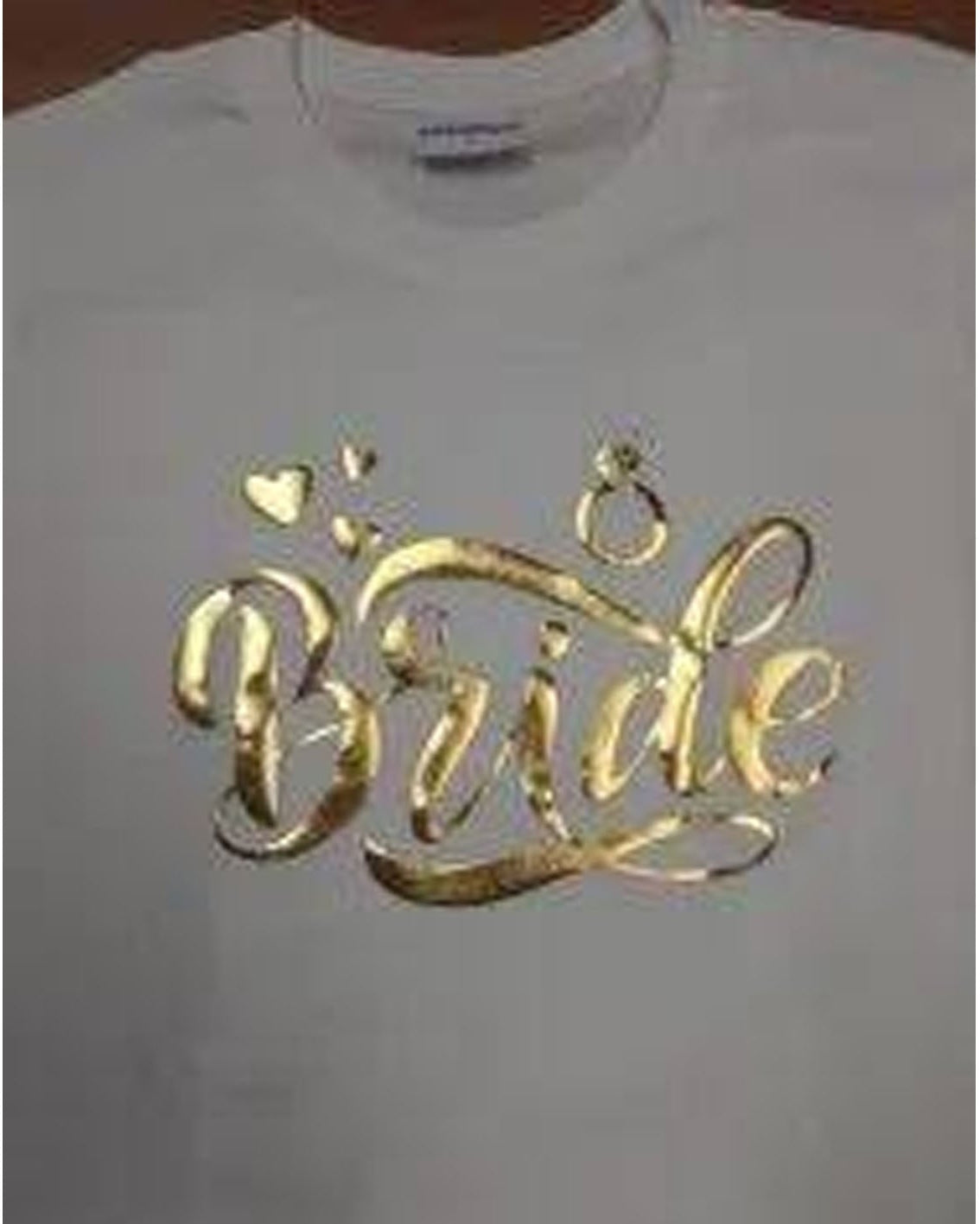 A white short-sleeve t-shirt featuring a metallic gold foil graphic that reads 'Bride' in cursive, decorated with small hearts and a ring motif, shown on a hanger.