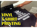 A black t-shirt with white printed text and a crown graphic is being held under a heat press, with large yellow text overlay reading 'VINYL T-SHIRT PRINTING'.